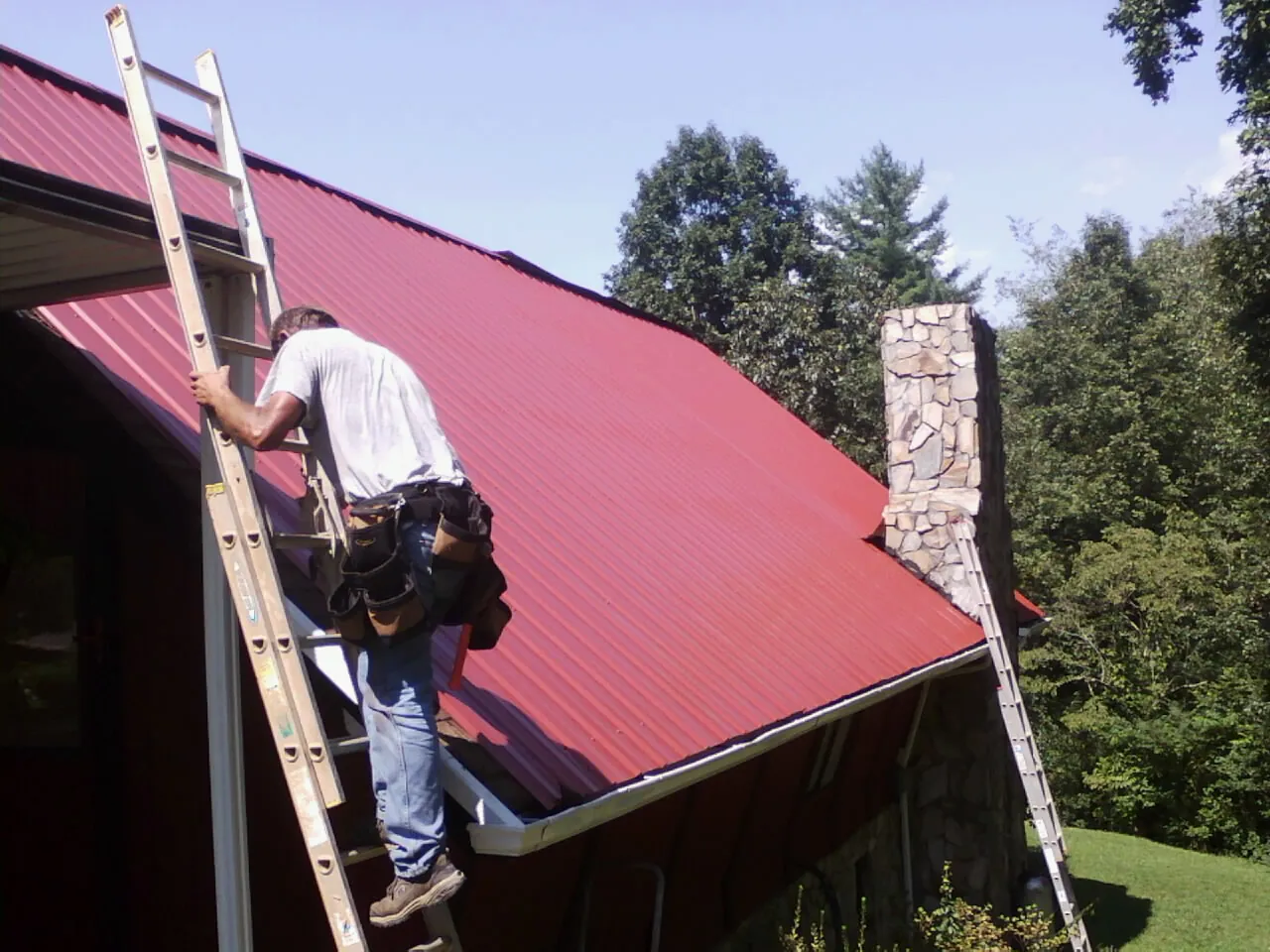 Roofer on a ladder beside a red metal roof and stone chimney — Soffit & Fascia Repair by Roofing Brothers in Whiteford